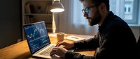 Young adult male with dark hair and beard, wearing glasses and dark clothing, analyzing stock market charts on laptop at wooden desk in modern office setting with warm pendant lighting and city view in background.