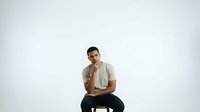 Young man with dark hair and olive complexion wearing casual beige t-shirt and black pants, sitting in contemplative pose against white background for portrait and lifestyle use.