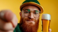Smiling bearded hipster man in green shirt and cap holding craft beer glass against yellow background, showing thumbs up gesture.