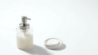 Modern glass soap dispenser with stainless steel pump and ceramic dish on white background for bathroom decor and hygiene products.