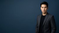 Young Asian male professional in dark blazer and black shirt against blue studio background, suitable for corporate portraits and business headshots.