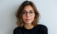 Young Caucasian woman with brown wavy bob haircut wearing glasses and black sweater smiling at camera against light wall, modern casual style portrait.