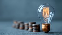 Glowing vintage light bulb with warm filament next to stacked coins on dark surface representing energy costs and electricity savings concept.