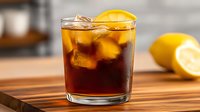 Glass of cold brew coffee with ice cubes and fresh lemon slice on wooden table, refreshing summer drink with citrus garnish and dark background.