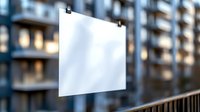 Blank white poster mockup hanging outdoors against urban apartment building background with bokeh lights and metal railing, ready for advertising design.
