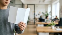 Young man in casual gray shirt holding blank white tri-fold brochure in modern office environment with colleagues working at desks in background, suitable for marketing and business communications.