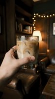 Hand holding a glass of iced coffee or cold beverage in a cozy interior setting with warm string lights and ambient lighting in the background.