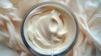 Luxurious white face cream or moisturizer in glass jar with silky smooth swirled texture, captured from above on soft fabric background for cosmetic and skincare marketing.