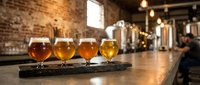 Craft beer flight tasting sampler with four different ales on wooden paddle in rustic brewery taproom with exposed brick walls and brewing equipment.