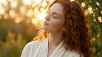 Young woman with curly red hair enjoying peaceful moment in golden sunlight outdoors with eyes closed, expressing serenity and mindfulness in nature.