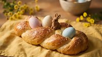 Easter braided bread with colorful eggs and chocolate bunny on wooden table with yellow flowers creating festive holiday atmosphere for spring celebration.