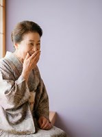 Mature Asian woman in traditional beige kimono laughing with hand near mouth against lavender background, expressing joy and happiness for lifestyle and cultural content.