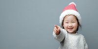 Joyful Asian child wearing Santa hat pointing at camera with excited expression against gray background, holiday season concept.