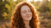 Young redhead woman with curly hair enjoying peaceful moment outdoors with eyes closed in warm golden sunlight creating serene natural portrait.