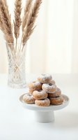 Fresh homemade donuts dusted with powdered sugar arranged on white ceramic cake stand with dried pampas grass in crystal vase creating elegant bakery display.