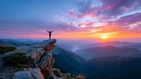 Silhouette of person with arms outstretched on mountain cliff edge against dramatic sunset sky over misty valley landscape.