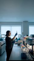 Young Hispanic female lab technician examines blood sample tube in modern clinical laboratory surrounded by colorful specimen racks.