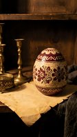 Ornate decorated Easter egg with traditional folk patterns sits on vintage wooden table with brass candlestick in warm candlelit atmosphere for holiday celebrations.