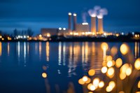 Blurred industrial power plant at night with glowing lights reflecting on water surface, creating dreamy bokeh effect in foreground. Atmospheric evening cityscape.