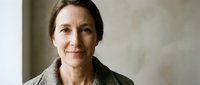 Middle-aged woman with brown hair, Caucasian ethnicity, wearing olive green jacket, neutral expression, studio portrait against beige wall background.