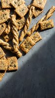 Assorted homemade crackers and breadsticks topped with sesame seeds and herbs arranged on gray concrete surface for food photography and culinary content.