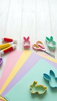 Colorful Easter craft supplies with bunny cookie cutters, scissors, glue sticks and pastel paper sheets on white wooden background for DIY projects.