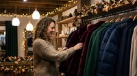 Man with long curly hair browsing winter coats in boutique clothing store with warm lighting and holiday decorations, shopping for seasonal outerwear.