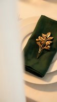 Elegant table setting with dark green folded napkin and gold leaf brooch on white plate, warm natural lighting for fine dining and event styling.
