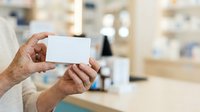 Senior male pharmacist holding blank white business card in modern pharmacy setting with blurred shelves and products in background.