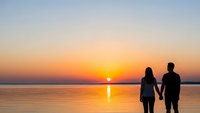 Romantic couple silhouettes holding hands watching golden sunset over calm water with warm orange and pink sky colors reflecting on lake surface.