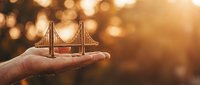 Hand holding miniature suspension bridge model against warm golden bokeh background, symbolizing infrastructure development, engineering innovation, and business growth concepts.