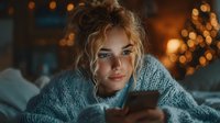 Young blonde woman lying in bed using smartphone with warm bokeh lights creating cozy evening atmosphere for social media and technology concepts.