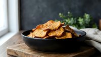 Crispy baked sweet potato chips in a dark bowl on a rustic wooden board, seasoned with herbs, natural light, healthy snack concept.