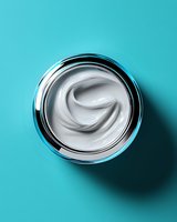White luxurious face cream in glass jar with metallic rim on turquoise background with soft shadow, top view. Cosmetic product photography for beauty industry.