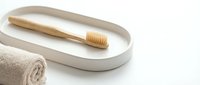 Bamboo toothbrush on a minimalist ceramic tray beside a rolled beige towel, eco-friendly bathroom essentials on white background.