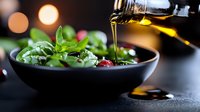 Olive oil being poured over fresh green salad with basil leaves and dried tomatoes in black ceramic bowl against dark background with bokeh lights.