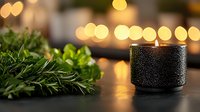 Black textured candle holder with burning flame against blurred golden bokeh lights, fresh green herbs on wooden surface creating cozy atmosphere.