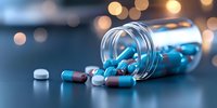 Blue and white capsule pills spilling from transparent prescription bottle on dark surface with soft bokeh lights creating medical atmosphere.
