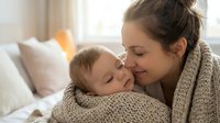 Young Caucasian mother in knit sweater tenderly kissing sleeping newborn baby wrapped in cozy blanket on bed. Intimate maternal bonding moment at home.