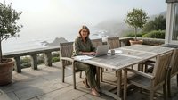Young professional woman working remotely on laptop at outdoor wooden table overlooking scenic coastal landscape with mountains and ocean views.
