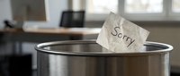 Crumpled paper note with the word Sorry thrown into a metal trash can in a blurred office interior, symbolizing regret and apology.