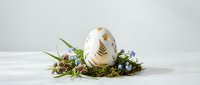 White Easter egg with golden feather pattern nestled in natural moss and spring flowers arrangement on clean background for holiday decorating.