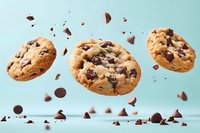 Three chocolate chip cookies floating with crumbs and chocolate pieces scattered around on light blue background for bakery advertising and food design.