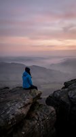 Woman in blue jacket sitting on rocky cliff overlooking misty valley landscape at sunrise or sunset. Contemplative outdoor scene for travel, wellness, and nature themes.