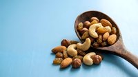 Wooden spoon with mixed nuts including almonds, cashews, walnuts and hazelnuts spilling onto blue surface for healthy snacking and cooking ingredients.