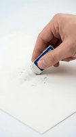Hand holding blue eraser removing pencil marks from white paper creating rubber shavings on clean surface for correction and editing work.
