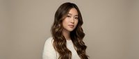 Young Asian woman with long wavy brown hair wearing cream sweater against neutral beige background for fashion and beauty content.