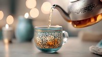 Steaming hot tea being poured into ornate turquoise glass cup with golden oriental pattern against blurred bokeh background, cozy atmospheric drink photography.