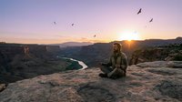 Man in outdoor gear sitting on rocky cliff overlooking winding river valley at sunset with flying birds and mountains in distance, adventure and nature exploration concept.