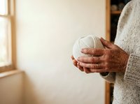 Senior man with brown skin holding white yarn ball, wearing gray sweater indoors near window. Concept for crafting, knitting hobby, leisure activities, retirement lifestyle, handmade projects.